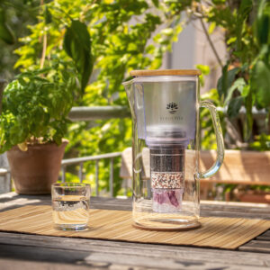 Eine Glas-Filterkanne für Wasser mit Holzdeckel steht neben einem gefüllten Wasserglas auf einem Tisch im Grünen.