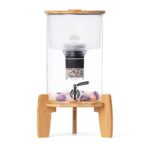 Ein Stand-Wasserfilter aus Glas mit einem Sockel und Deckel aus Bambus, einem Edelstahl-Zapfhahn und Edelsteinen im Behälter.