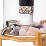 Detailansicht eines Glas-Wasserfilters mit mehrstufiger Filterkartusche und Edelsteinen wie Rosenquarz und Amethyst, stehend auf einem Bambussockel.