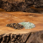 Zwei kleine Haufen getrommelter Edelsteine, Fluorit und Tigerauge, liegen auf einer rustikalen Holzoberfläche.