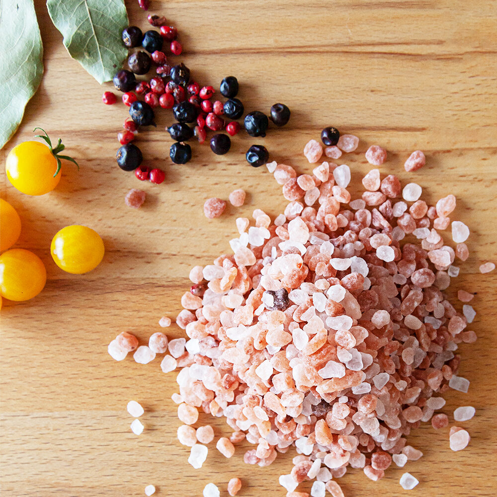 Grobkörniges Urmeer-Kristallsalz, arrangiert auf einem Holzbrett mit bunten Pfefferkörnern, Lorbeerblättern und gelben Kirschtomaten.