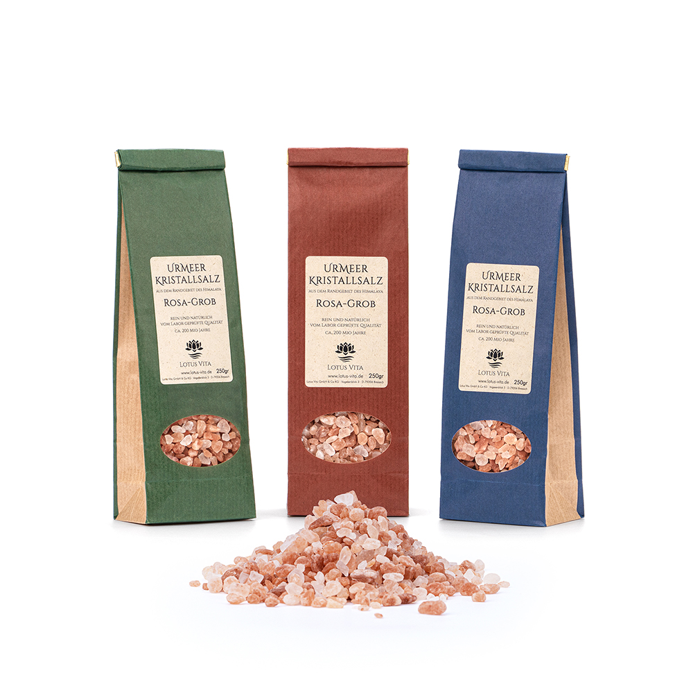 Drei verschiedenfarbige 250g-Packungen grobkörniges Urmeer-Kristallsalz von Lotus Vita, mit einem Haufen rosa Salzkristalle im Vordergrund auf weißem Hintergrund.