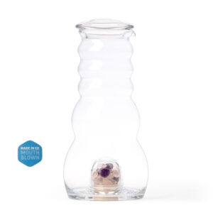 Mundgeblasene Glaskaraffe in wellenförmigem Design, mit Glasdeckel und einem integrierten Bodenfach, gefüllt mit einer bunten Edelsteinmischung.