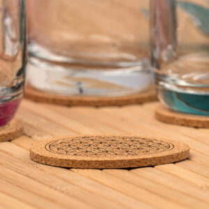 Ein runder Kork-Untersetzer mit eingravierter Blume des Lebens liegt auf einem Holztisch, während im unscharfen Hintergrund Wassergläser auf weiteren Untersetzern stehen.