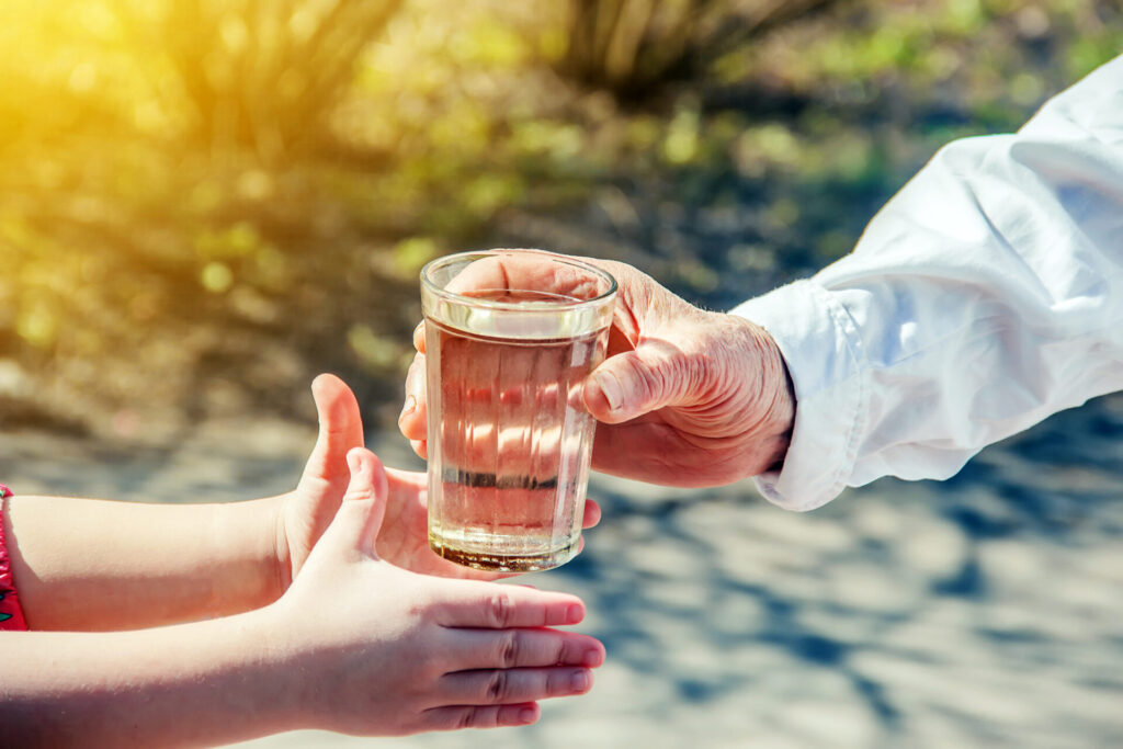 Die Hand einer älteren Person reicht einem Kind ein Glas mit klarem, reinem Wasser in einer sonnigen, natürlichen Umgebung.