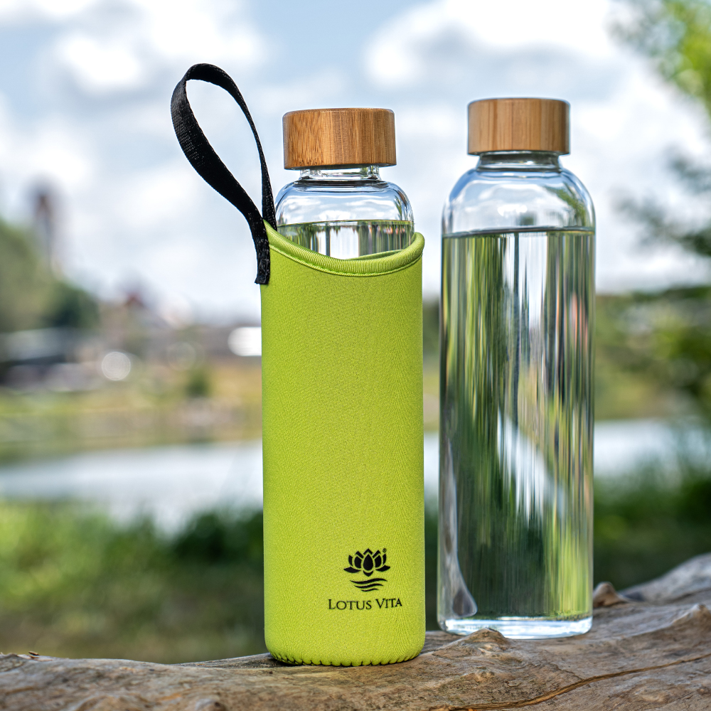 Zwei Glas-Trinkflaschen mit Bambusdeckel, eine davon in einer leuchtend grünen Neopren-Schutzhülle, stehen auf einem Baumstamm in der Natur.
