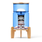 Eleganter Stand-Wasserfilter mit blauem Glasbehälter, Deckel und Sockel aus Bambusholz. Im Inneren sind eine mehrstufige Filterkartusche und Edelsteine zur Wasserveredelung sichtbar.