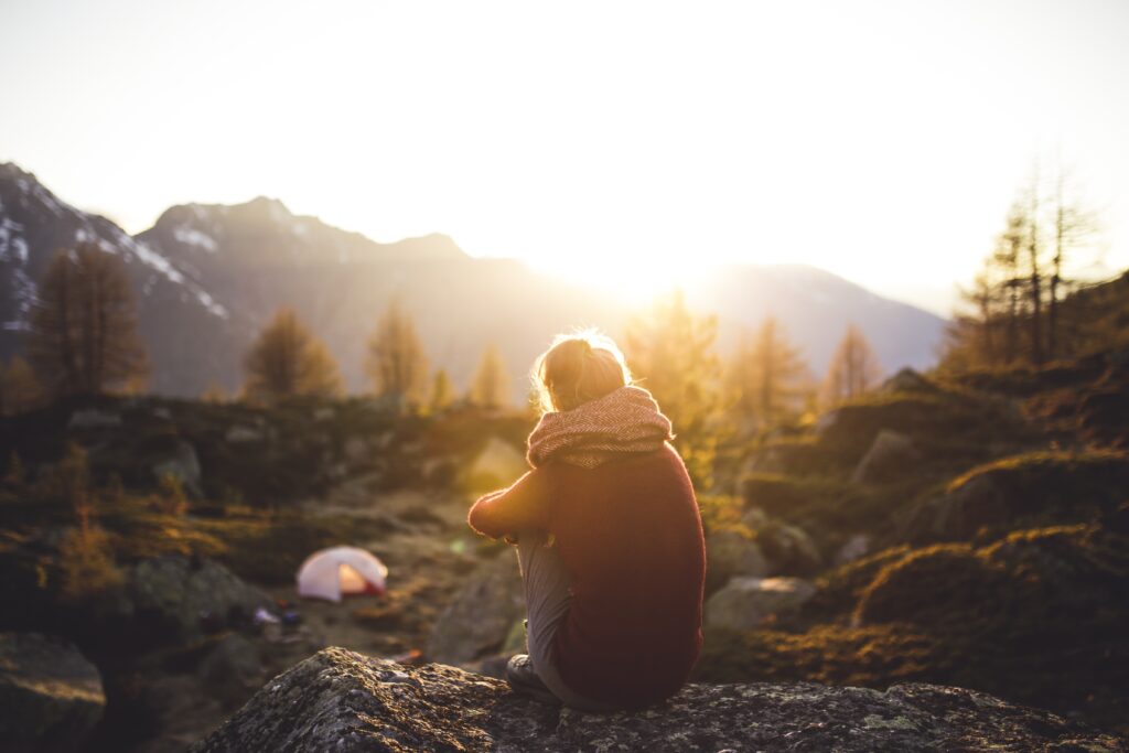 Eine Person sitzt bei Sonnenaufgang auf einem Felsen und blickt über eine weite Berglandschaft, ein Symbol für Freiheit und Unabhängigkeit auf Reisen.