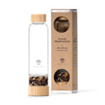 Eine Glas-Trinkflasche von Lotus Vita mit Bambusdetails und einem separaten Fach gefüllt mit Edelsteinen, stehend neben der Produktverpackung.