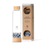 Eine Glas-Trinkflasche von Lotus Vita mit Bambusdeckel und einem separaten Fach gefüllt mit blauen und weißen Kristallen, neben der Produktverpackung aus Karton.