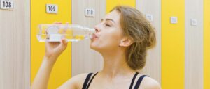 Eine sportliche Frau trinkt nach dem Training im Umkleideraum aus einer wiederverwendbaren Flasche, um ihren Körper zu rehydrieren.