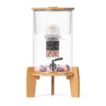 Ein Glas-Wasserspender von Lotus Vita auf einem Bambussockel, mit sichtbarer Filterkartusche und Edelsteinen im Inneren.