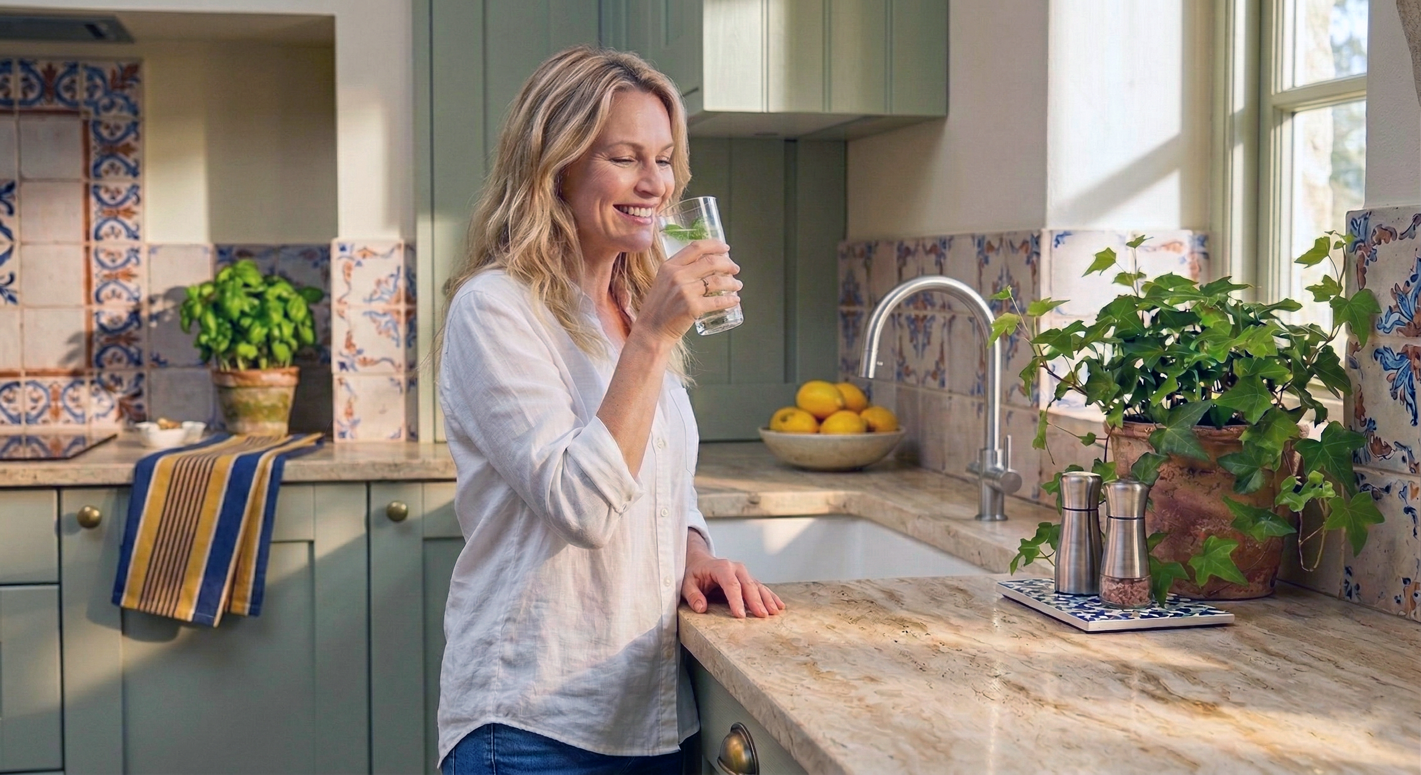 Eine blonde Frau steht in einer hellen, freundlichen Küche und trinkt lächelnd ein Glas klares Wasser.