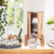 Ein Wasserfilter-Standgerät aus Akazienholz mit Glasbehälter, aufgestellt auf einer Theke in einem hellen Büroraum.